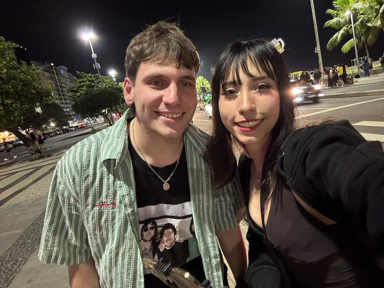 Gaspi con camisa de rayas verde y negro sonríe junto a un amigo de prenda negra en una calle iluminada.
