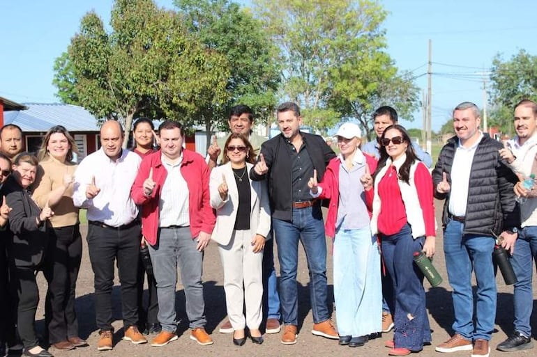 Grupo de 13 personas sonriendo y levantando el dedo índice en un evento al aire libre, con árboles y un edificio rojo de fondo.
