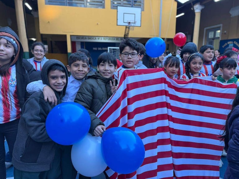 Grupo de niños sonriendo, vestidos con camisetas rojo y blanco, sosteniendo globos, en un ambiente festivo escolar.