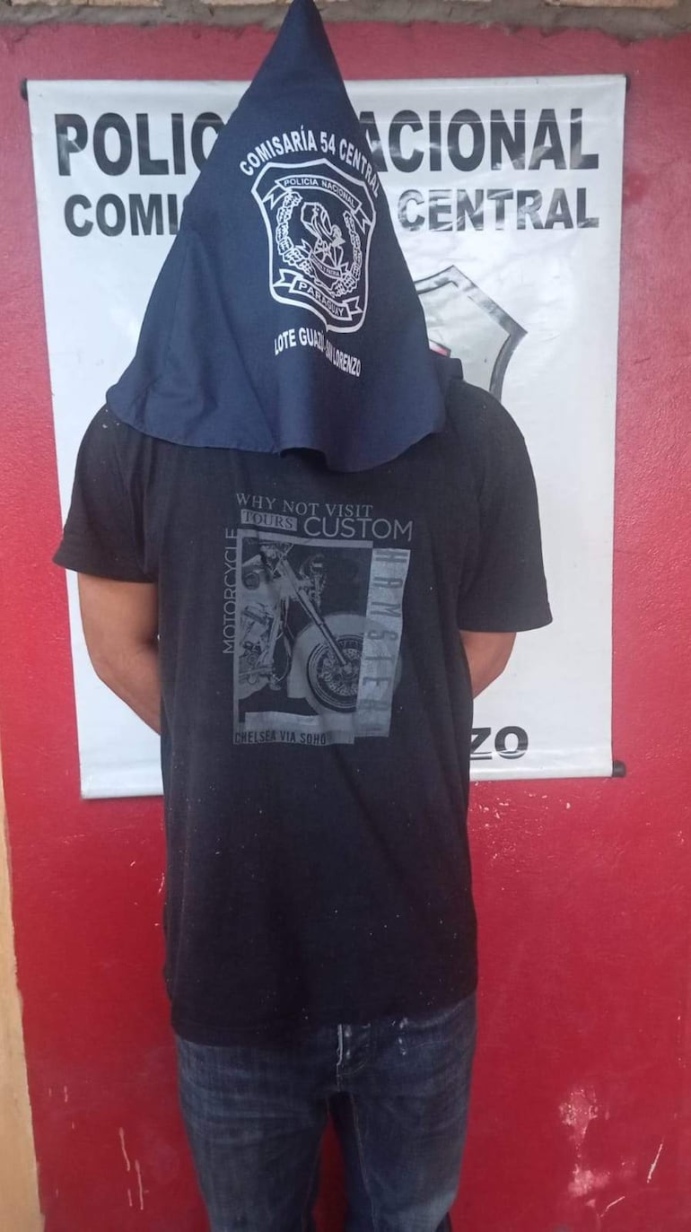 Hombre de espaldas con capuchón azul oscuro frente a la comisaría, en un ambiente rojo, esperando identificación.