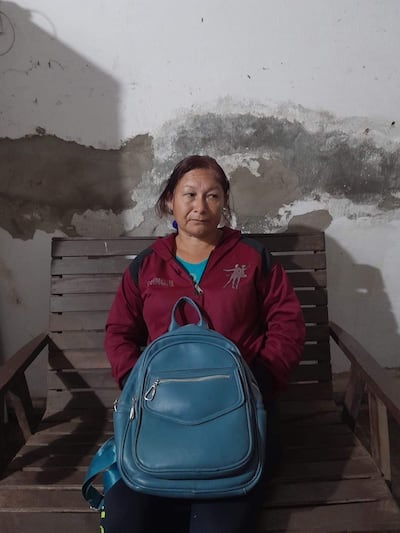 Mujer sentada en banco de madera, con chaqueta roja, camiseta azul y mochila a su lado, en un ambiente interior claro.