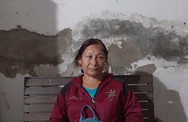 Mujer sentada en banco de madera, con chaqueta roja, camiseta azul y mochila a su lado, en un ambiente interior claro.