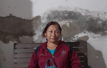 Mujer sentada en banco de madera, con chaqueta roja, camiseta azul y mochila a su lado, en un ambiente interior claro.