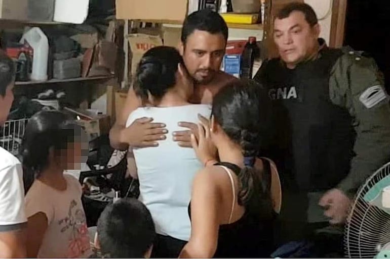 Morenita Marín semi-desnudo abrazado por una mujer con gesto afectuoso, mientras un hombre de la GNA observa y dos niñas miran atentas.