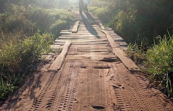 Persona con sombrero camina por un camino de madera, acompañada de una motocicleta, en un entorno rural con vegetación densa.