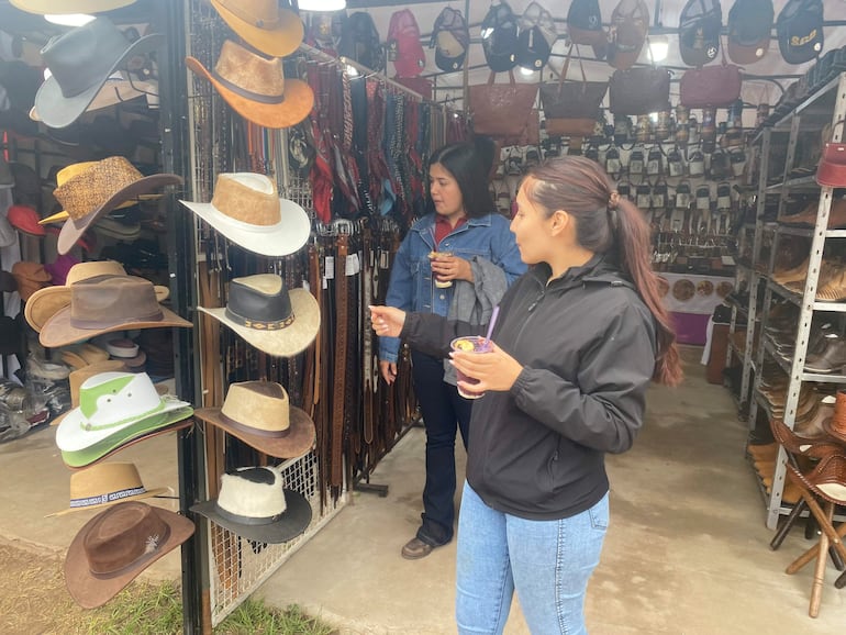 Dos mujeres en el mercado de la Expo Concepción 2026, una sostiene un vaso y otra mira con los brazos cruzados, rodeadas de sombreros de diferentes estilos.
