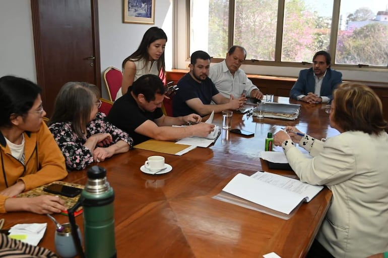 Mujer de pie con teléfono presenta información a grupo de ocho personas sentadas, en un ambiente iluminado y acogedor.