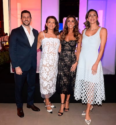 Cuatro mujeres en vestidos elegantes y un hombre en traje oscuro sonríen en un salón con luces moradas y naranjas.