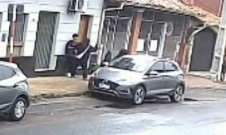 Dos personas, una con camiseta blanca y chaqueta oscura, conversan junto a un edificio en un ambiente urbano.