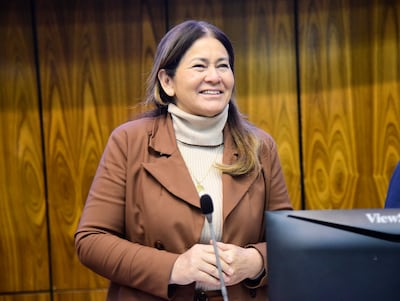 Mujer de pie sonriendo, con abrigo marrón y suéter beige, en un entorno formal con fondo de madera clara.