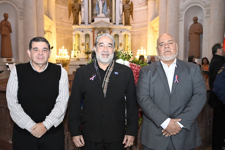 Tres hombres en una iglesia: uno con suéter negro, otro con abrigo y pañuelo, y el tercero con camisa blanca y saco gris, todos con cinta tricolor.