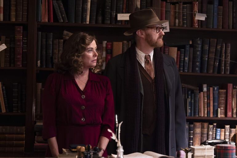 Mark Gatiss con sombrero y abrigo oscuro observa, mientras una mujer de vestido rojo se muestra serena en un entorno de estantes de libros.