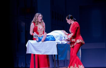 Lorna Cepeda en vestido rojo sostiene un número 69, mientras Natalia Ramírez observa un cuerpo cubierto en una mesa quirúrgica.