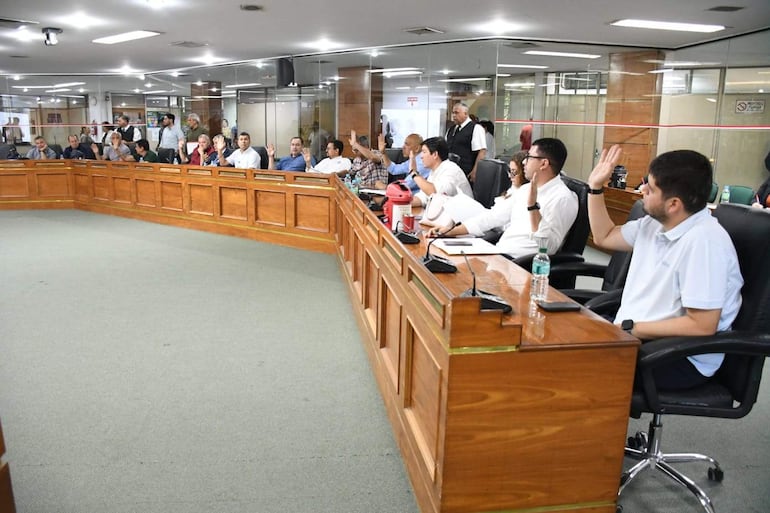 Javier Pintos, Jesús Lara y otros presentes con gestos tranquilos en un ambiente formal, levantando manos en votación.