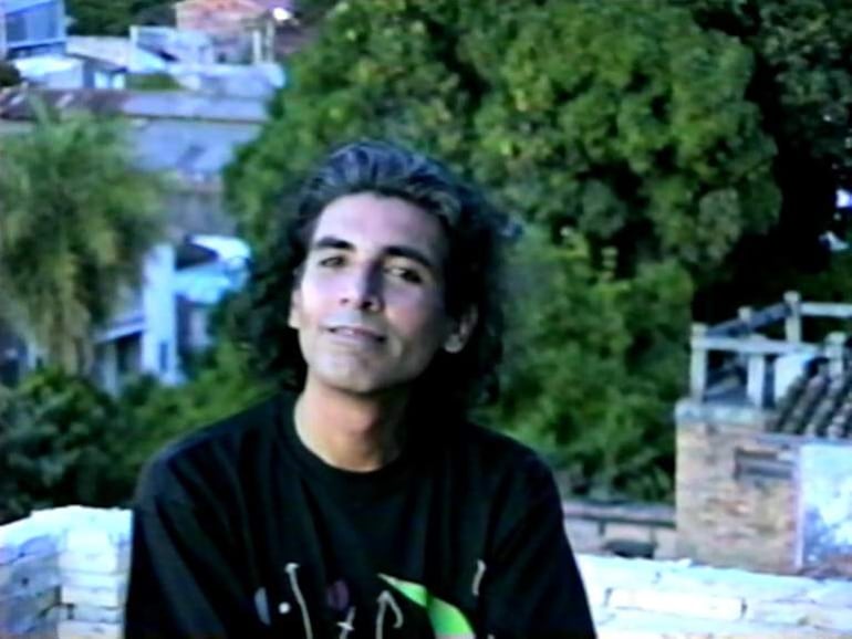 Un joven con cabello largo y oscuro, vestido con camiseta negra, sentado en un entorno relajado con edificaciones y árboles al fondo.
