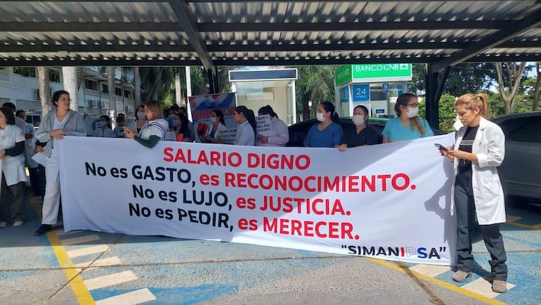 Grupo de médicos con uniformes y mascarillas sostienen pancarta sobre salario digno en ambiente exterior.