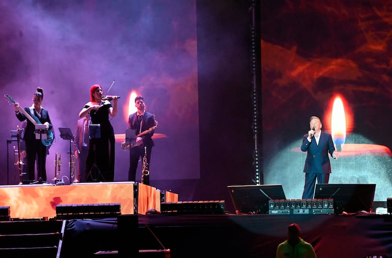 Montaner en traje oscuro y camisa blanca, cantando en un escenario iluminado; músicos acompañan tocando varios instrumentos.