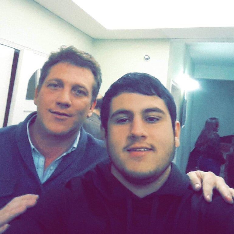 Acosta Vázquez sonríe con camiseta oscura y barba, junto a Domínguez, ambos en un ambiente informal con luz suave.