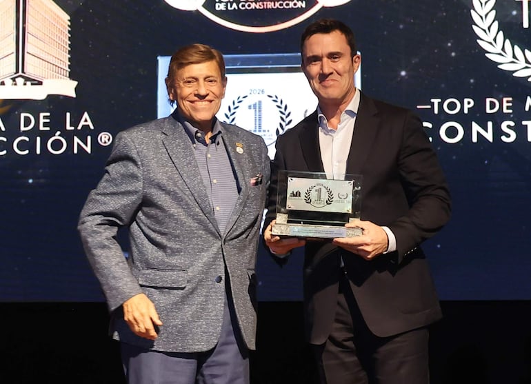 Dos hombres en gala: uno con chaqueta azul claro sonríe, mientras otro en traje oscuro sostiene un trofeo ante un fondo de logotipos.