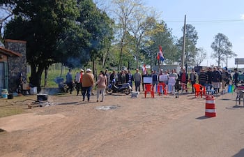 Grupo de personas sentadas y de pie en manifestación, algunas con sombreros, sosteniendo carteles y banderas paraguayas en un ambiente rural.