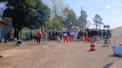 Grupo de personas sentadas y de pie en manifestación, algunas con sombreros, sosteniendo carteles y banderas paraguayas en un ambiente rural.