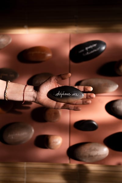 Mano sostiene una piedra ovalada con inscripcióm 'déjame ser', rodeada de otras piedras en un estudio iluminado.