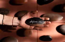 Mano sostiene una piedra ovalada con inscripcióm 'déjame ser', rodeada de otras piedras en un estudio iluminado.