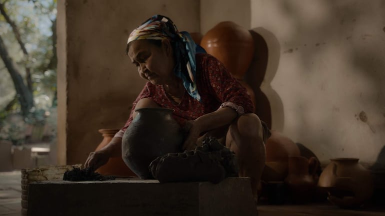 Mujer de edad avanzada, sentada en el suelo, vestida con blusa roja y pañuelo, modelando cerámica en un taller con paredes de barro.