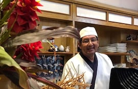 Diosnel Miranda sonriente, vestido con chaqueta blanca y delantal negro, de pie detrás de la barra en un restaurante decorado con flores.