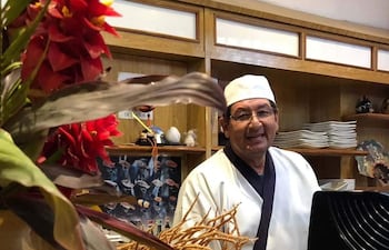 Diosnel Miranda sonriente, vestido con chaqueta blanca y delantal negro, de pie detrás de la barra en un restaurante decorado con flores.