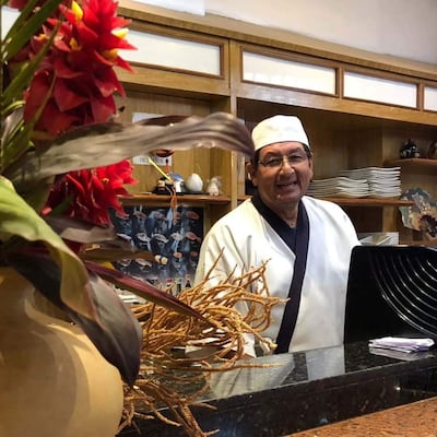 Diosnel Miranda sonriente, vestido con chaqueta blanca y delantal negro, de pie detrás de la barra en un restaurante decorado con flores.