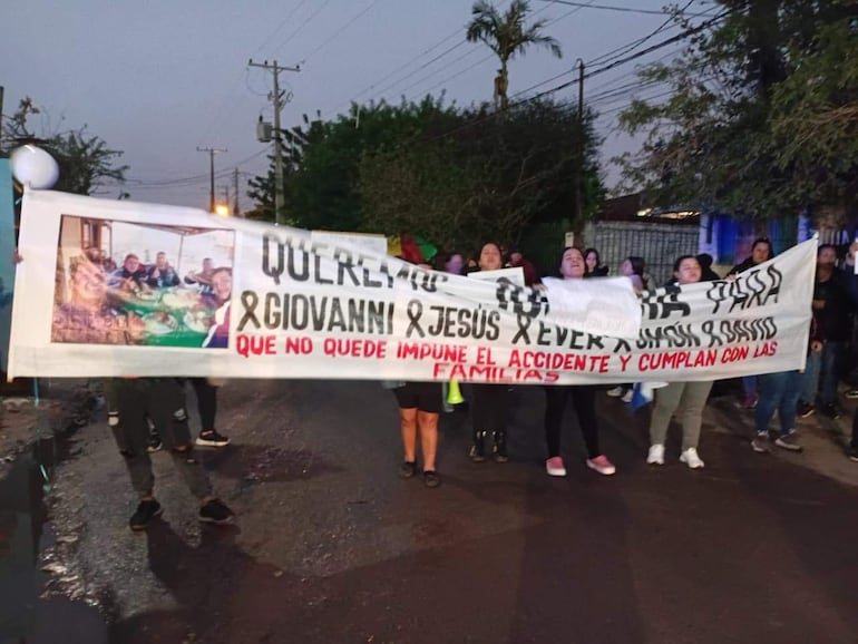 Grupo de diez personas sosteniendo un gran cartel con nombres y demanda de justicia, expresiones activas y ambiente urbano.