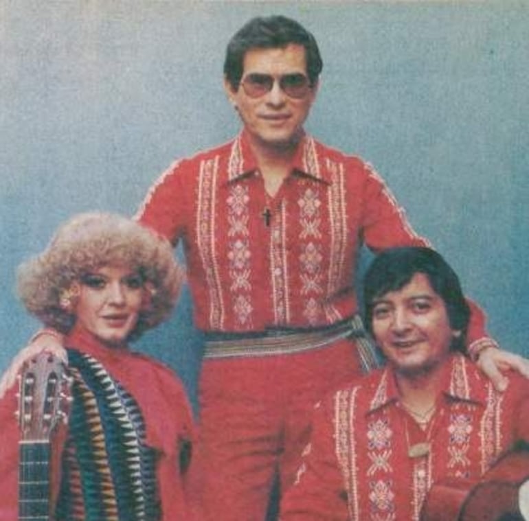 Tres hombres con camisas rojas, uno con gafas oscuras, posan con guitarra y guitarra acústica en un ambiente amigable.