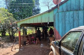 Varias personas conversan bajo un toldo en un ambiente rural, con un vehículo estacionado y árboles alrededor.