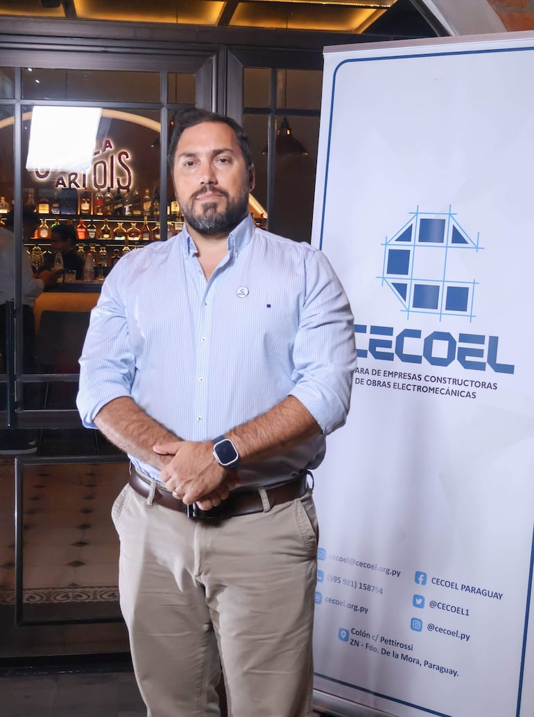 Hombre de pie con expresión seria, camisa de rayas azul y reloj, frente a banner de CECOEL en un bar iluminado.
