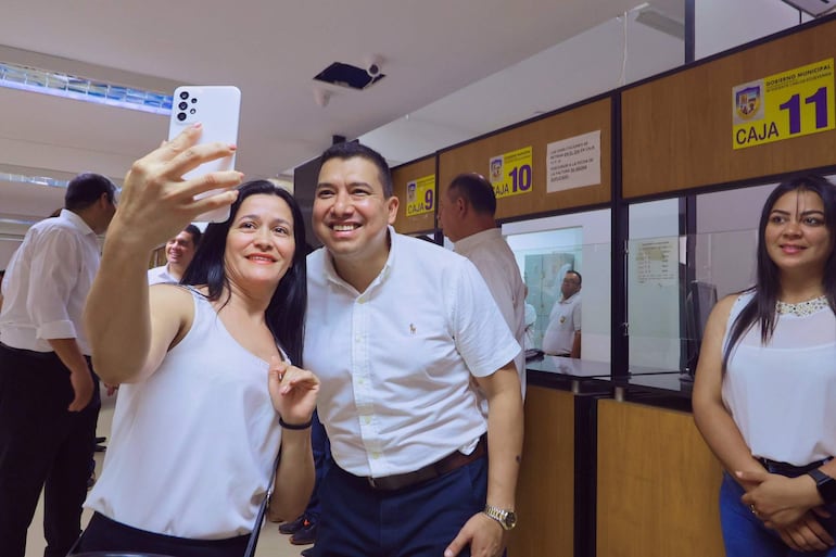 Mujer de cabello largo y oscuro sonríe mientras toma una selfie con un hombre en camisa blanca, en un ambiente de oficina.