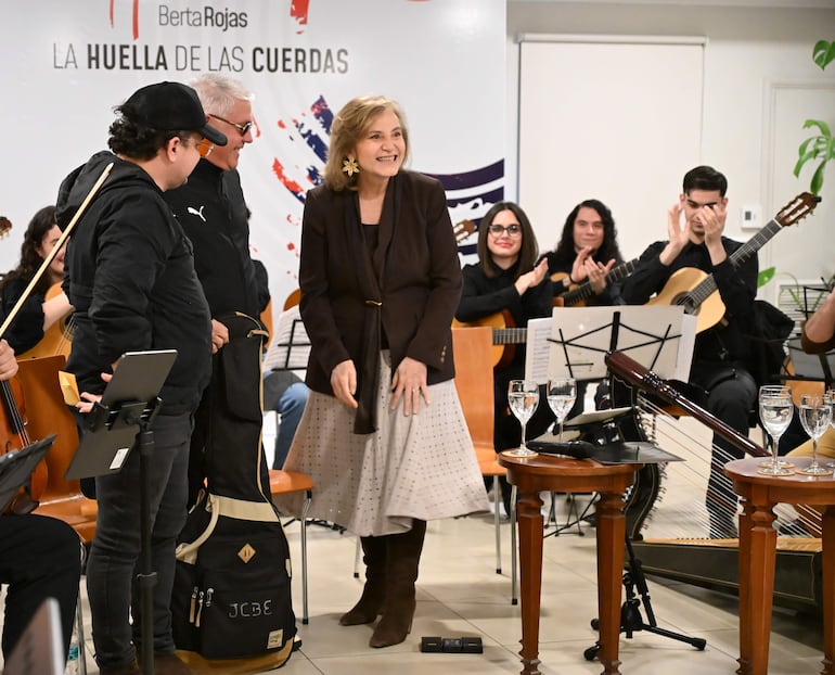Berta Rojas sonriente con blusa marrón y falda clara, rodeada de músicos en evento musical en ambiente festivo.