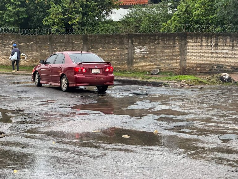 Hombre con capucha y bolsa camina por acera mientras un sedán rojo atraviesa una calle llena de baches y agua.