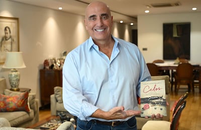Carlos Mateo Balmelli sonriente en camisa celeste, sosteniendo su libro 'Celos' en un ambiente iluminado con muebles decorativos.