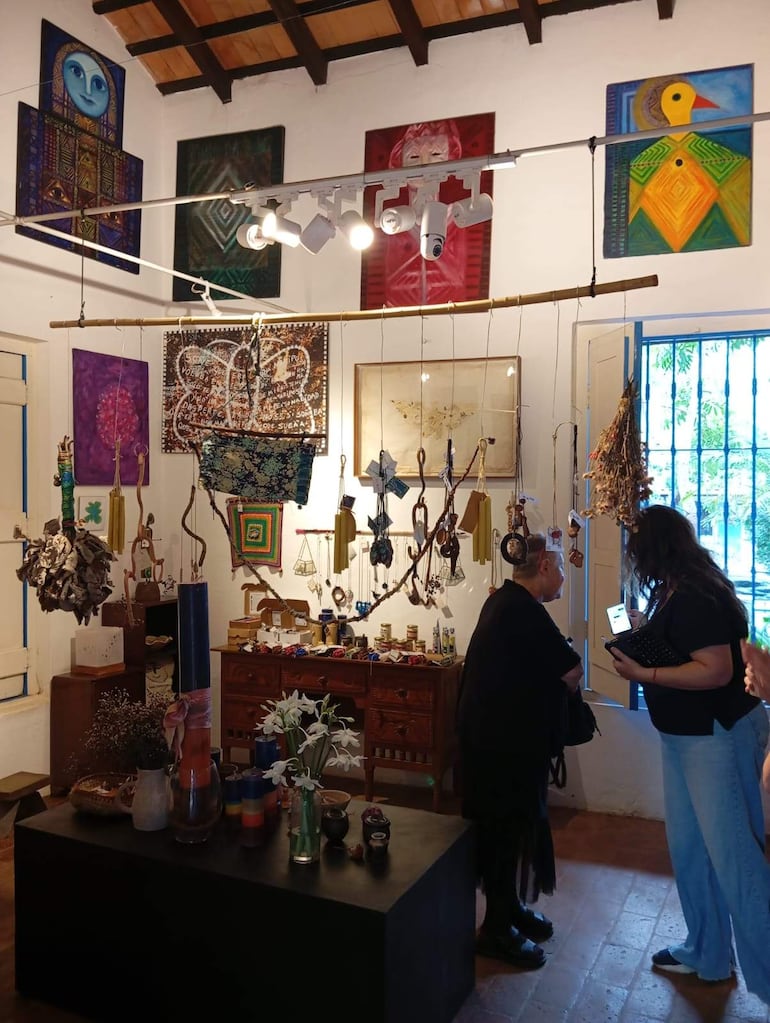 Mujeres en galería, una de perfil mirando una ventana, otra con móvil, rodeadas de obras de arte y flores blancas.