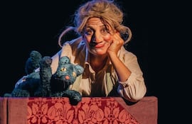 Mujer con maquillaje teatral y cabello despeinado, sentada en una mesa con un muñeco de gato de peluche verde.