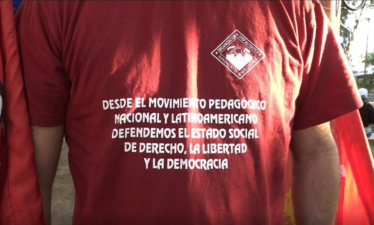 Docente de espaldas con camiseta roja que dice 'defendemos el estado social de derecho, la libertad y la democracia' en evento al aire libre.