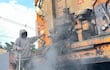 Trabajador de la construcción con sudadera y capucha, opera máquina de asfalto en una calle con casas de ladrillo al fondo.