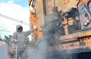 Trabajador de la construcción con sudadera y capucha, opera máquina de asfalto en una calle con casas de ladrillo al fondo.