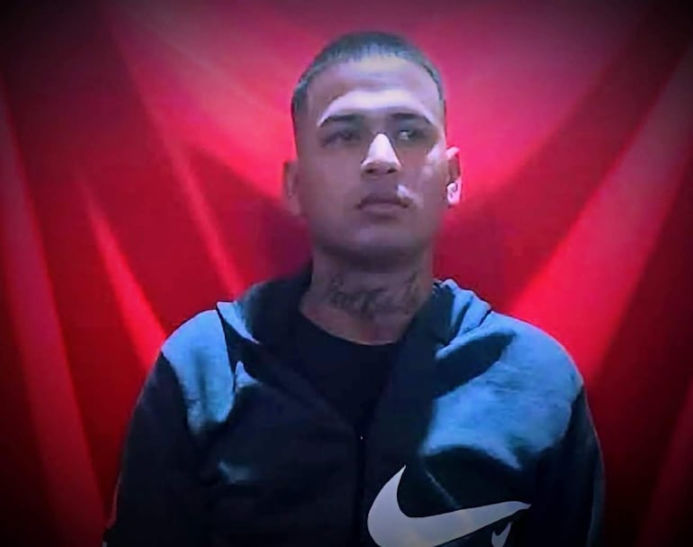 Hombre de cabello rapado y tatuaje en el cuello, vestido con hoodie oscuro y fondo rojo brillante.