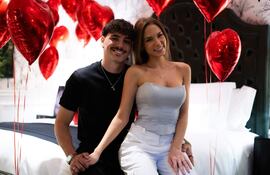 Hombre con cabello rizado y camiseta negra sonríe junto a una mujer en blusa gris y pantalones blancos, rodeados de globos rojos.
