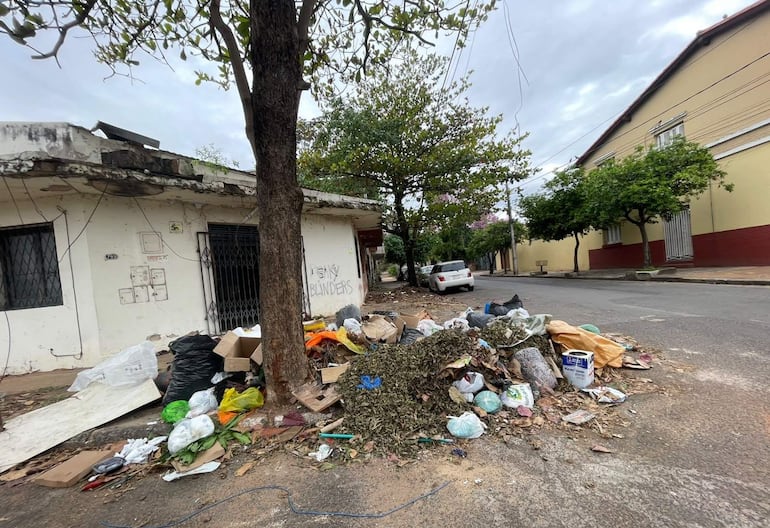 Basura variada en calle urbana desordenada, con casa blanca y vehículo estacionado a la derecha.
