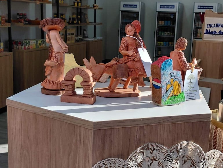 Artesanías de barro con figuras tradicionales, exhibidas sobre mesa blanca en un entorno iluminado, en tienda de productos locales.