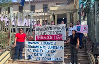 Grupo de manifestantes sosteniendo carteles, mujer con camiseta roja y hombre de camisa blanca enérgicos frente a edificio.