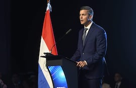 Santiago Peña, con traje oscuro y camisa blanca, habla desde un atril frente a una bandera de Paraguay en un auditorio.
